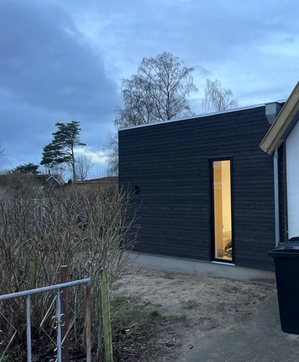 Tilbygning med sort træbeklædning og højt vinduesparti i aftenlys
