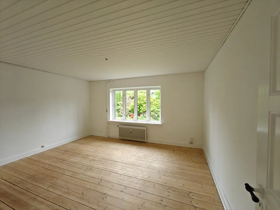 Tomt værelse med hvide vægge, hvidt loft af profilbrædder, lyst plankegulv og et vindue med fire fag og grønt udenfor