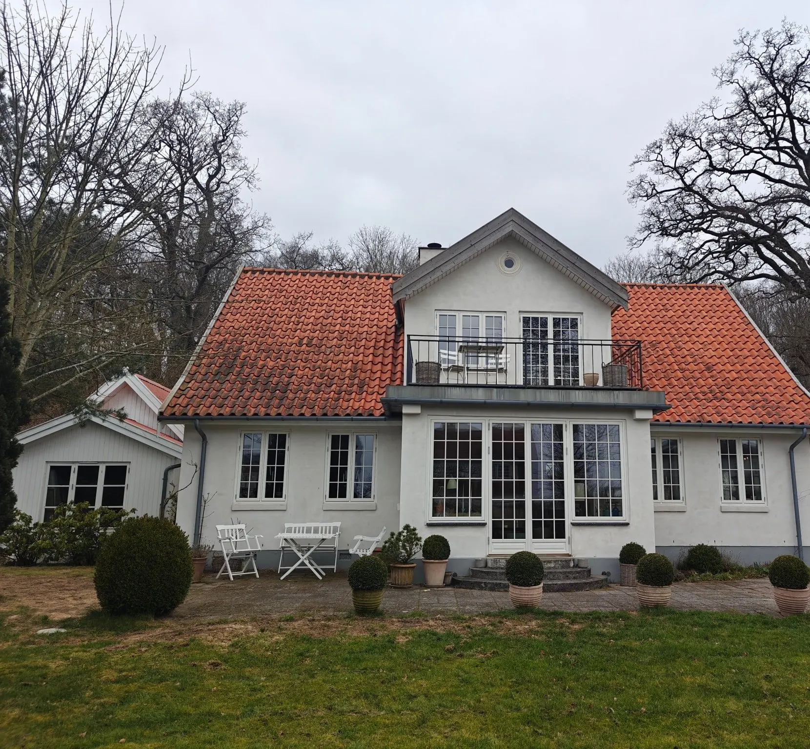 Hvidpudset villa med rødt tegltag set fra haven en grå dag, før malerarbejdet: grå vindskeder på gavlen, hvide sprossede vinduer og en lav sidebygning med hvid, lodret bræddebeklædning