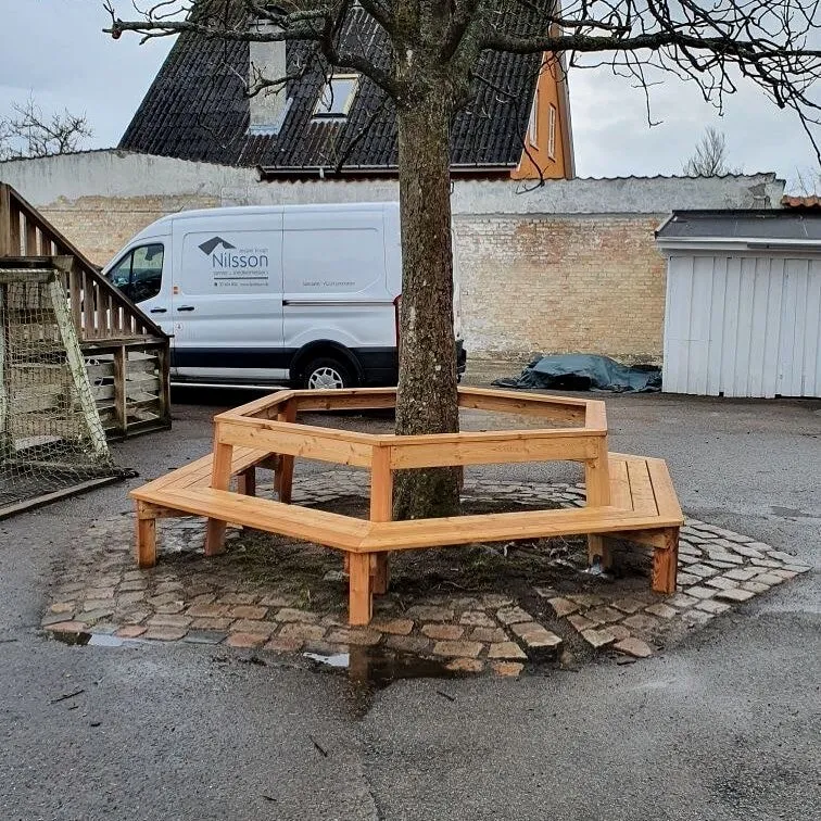 Håndlavet sekskantet træbænk bygget omkring et træ, med firmabilen i baggrunden