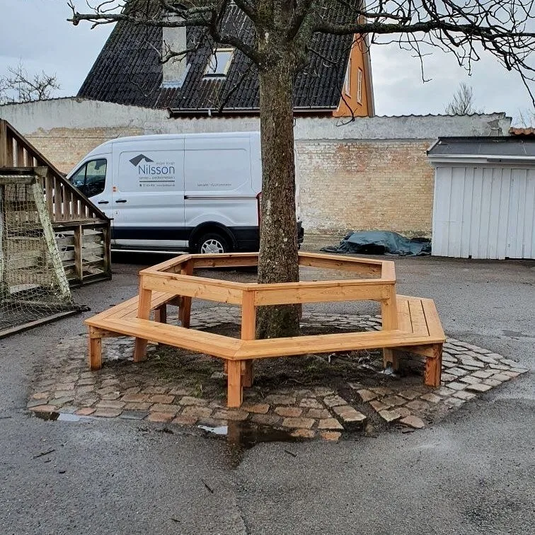 Håndlavet sekskantet træbænk bygget omkring et træ, med firmabilen i baggrunden